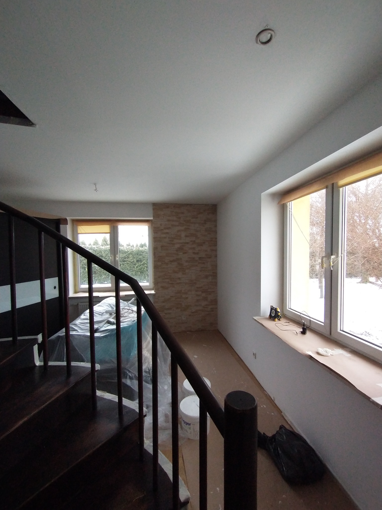 Pokój w trakcie remontu: zabezpieczone meble folią, widoczne wiaderka z farbą, świeżo pomalowane ściany, drewniane schody z brązową balustradą, okno z widokiem na zimowy krajobraz.