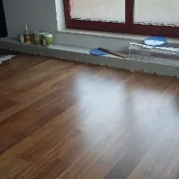 Częściowo ułożona, błyszcząca podłoga z paneli drewnopodobnych. Przy ścianie, na listwie przypodłogowej, widoczne pojemniki z farbą i akcesoria montażowe.