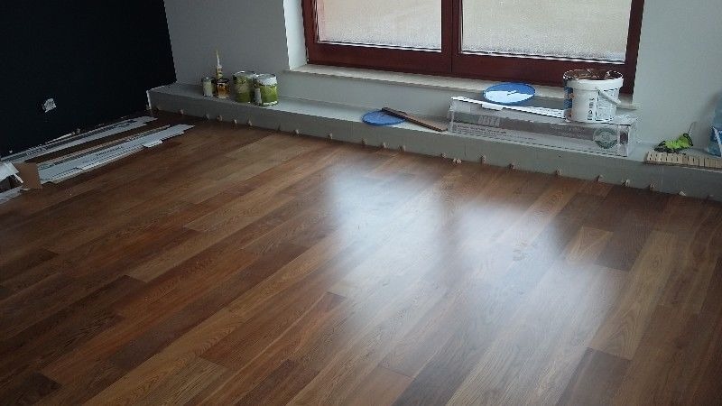 Częściowo ułożona, błyszcząca podłoga z paneli drewnopodobnych. Przy ścianie, na listwie przypodłogowej, widoczne pojemniki z farbą i akcesoria montażowe.