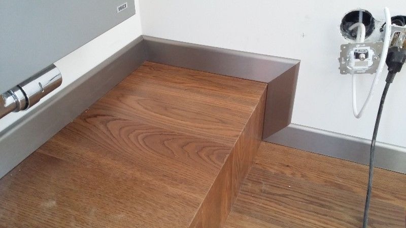 Wykończony fragment podłogi z drewnopodobnych paneli, z widoczną listwą przypodłogową w kolorze szarym oraz fragmentem grzejnika i gniazdkiem elektrycznym w ścianie.