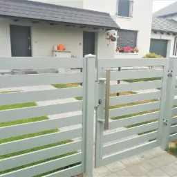 Nowoczesna, pozioma brama panelowa w kolorze szarym z metalowym uchwytem, widok z zewnątrz na posesję z kostki brukowej i dom z szarą stolarką okienną.