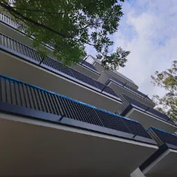 Nowoczesne balkony z czarnymi, metalowymi balustradami w nowym budynku wielorodzinnym, częściowo zabezpieczone folią ochronną, widok z dołu na tle błękitnego nieba z chmurami i zieleni drzew.