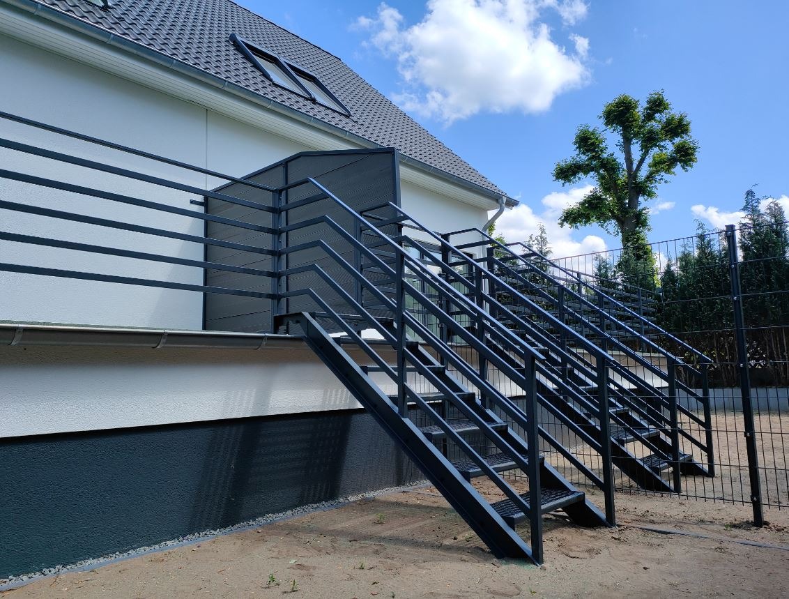 Zewnętrzne schody metalowe z balustradą prowadzące do budynku z białymi ścianami i ciemnym dachem, widoczne drzewa i ogrodzenie siatkowe w tle.