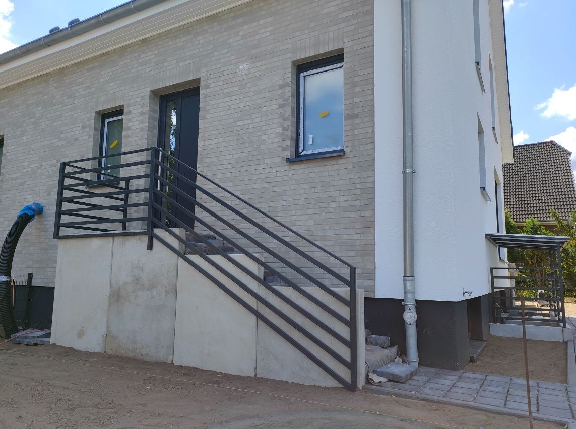 Nowoczesna, ciemnoszara balustrada schodowa z poziomych profili zamontowana przy wejściu do domu z szarej cegły licówki.