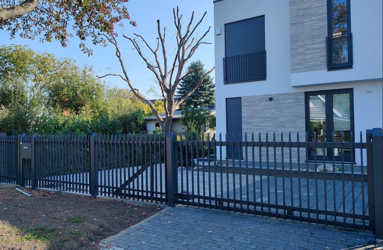 Nowoczesne ogrodzenie metalowe z pionowymi prętami w kolorze grafitowym, zintegrowane ze skrzynką na listy, na tle domu z szarej cegły i drzewa bez liści.