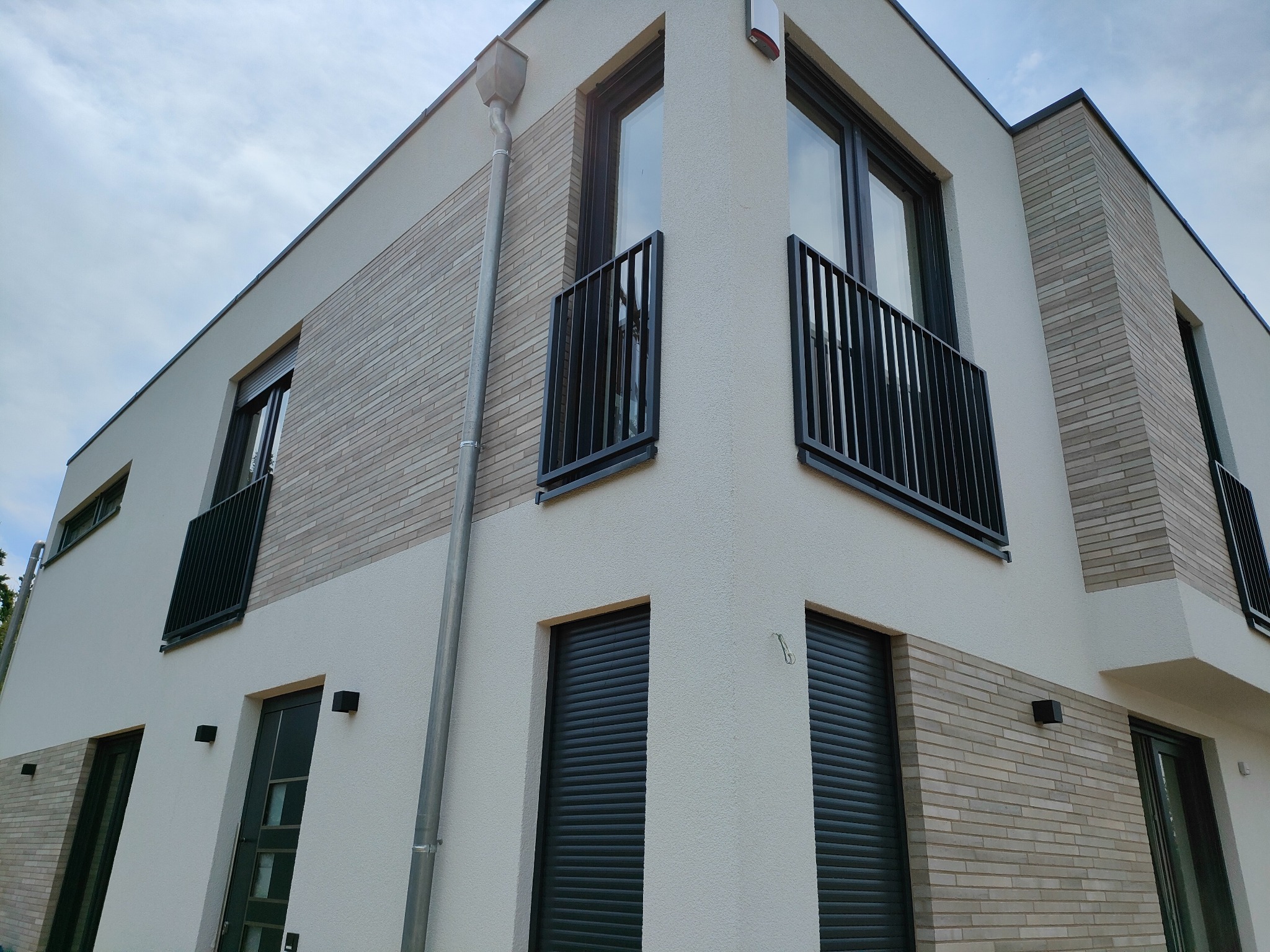 Nowoczesny, dwupiętrowy dom z jasną elewacją, ceglanymi akcentami i czarnymi balustradami balkonowymi, widziany z dołu pod kątem.