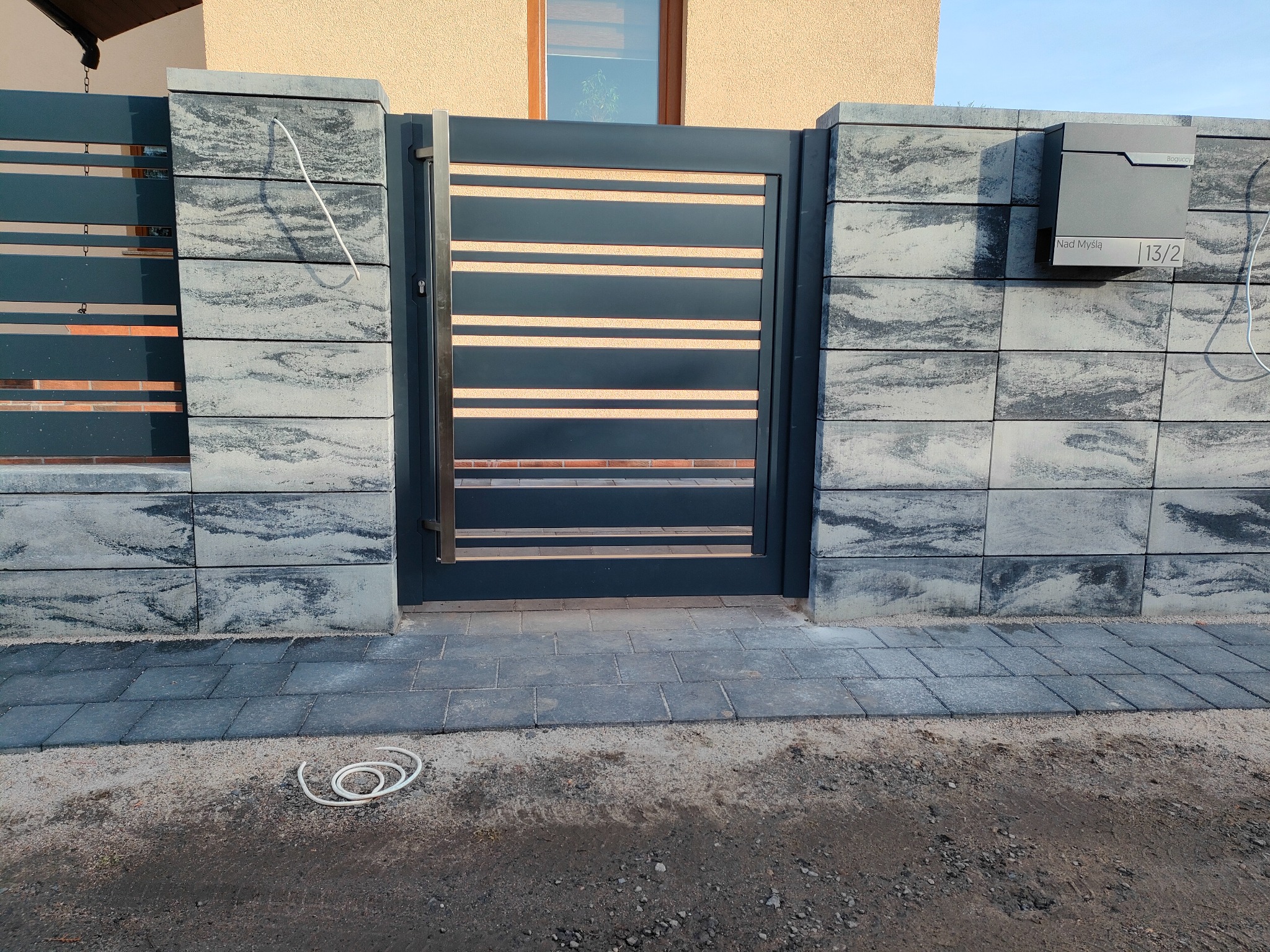 Nowoczesna brama wjazdowa z poziomymi, drewnopodobnymi panelami, osadzona w murze z szarych, cieniowanych bloczków betonowych; skrzynka pocztowa przymocowana do muru, kabel leżący na ziemi.