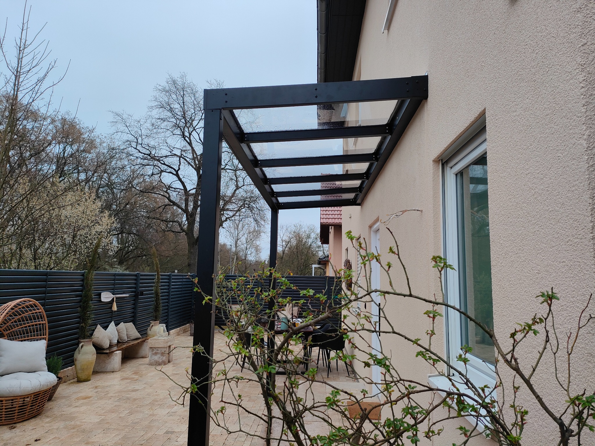 Czarna, nowoczesna pergola z przezroczystym zadaszeniem przylegająca do jasnej ściany budynku, z widocznym fragmentem tarasu z meblami ogrodowymi i dekoracyjnym ogrodzeniem w tle.