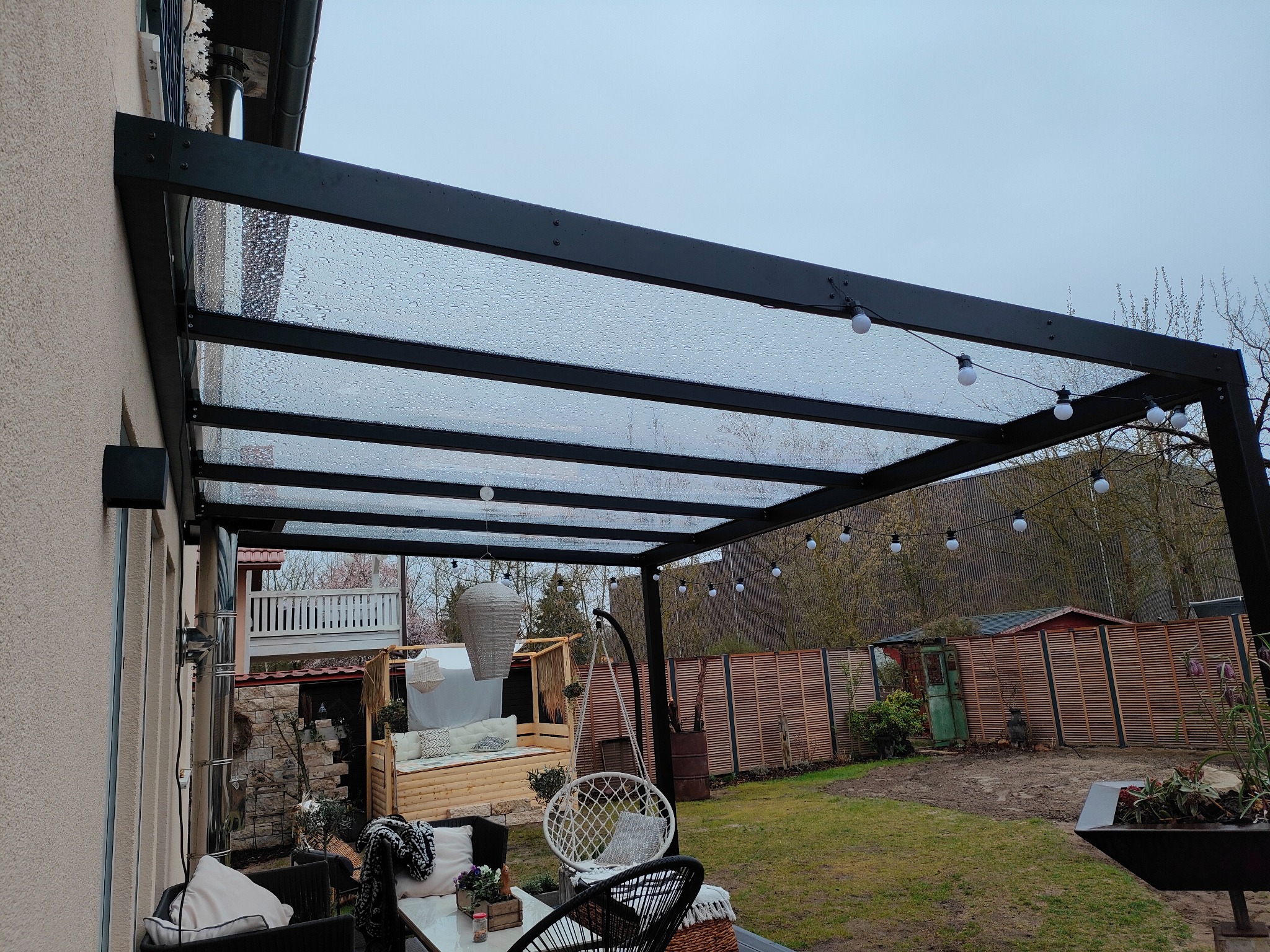 Zadaszenie tarasu z czarnej stali i przezroczystego poliwęglanu w Dębnie, oświetlone girlandą żarówkową, widoczne krople deszczu na powierzchni.