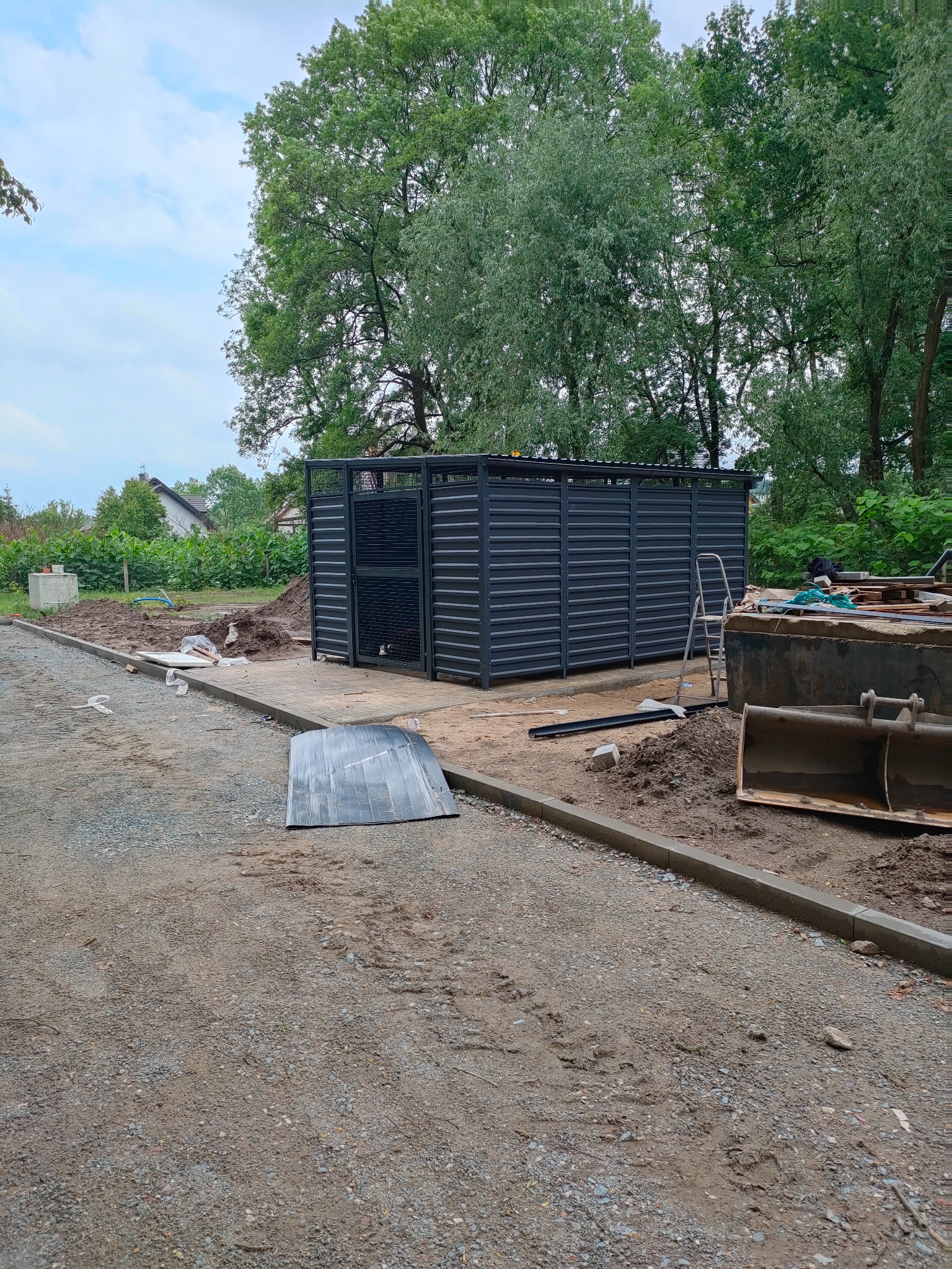 Nowoczesna, grafitowa konstrukcja osłonowa z metalowych paneli, prawdopodobnie na pojemniki na śmieci, z elementami ażurowymi, widoczna na terenie budowy z rozkopaną ziemią i sprzętem budowlanym...