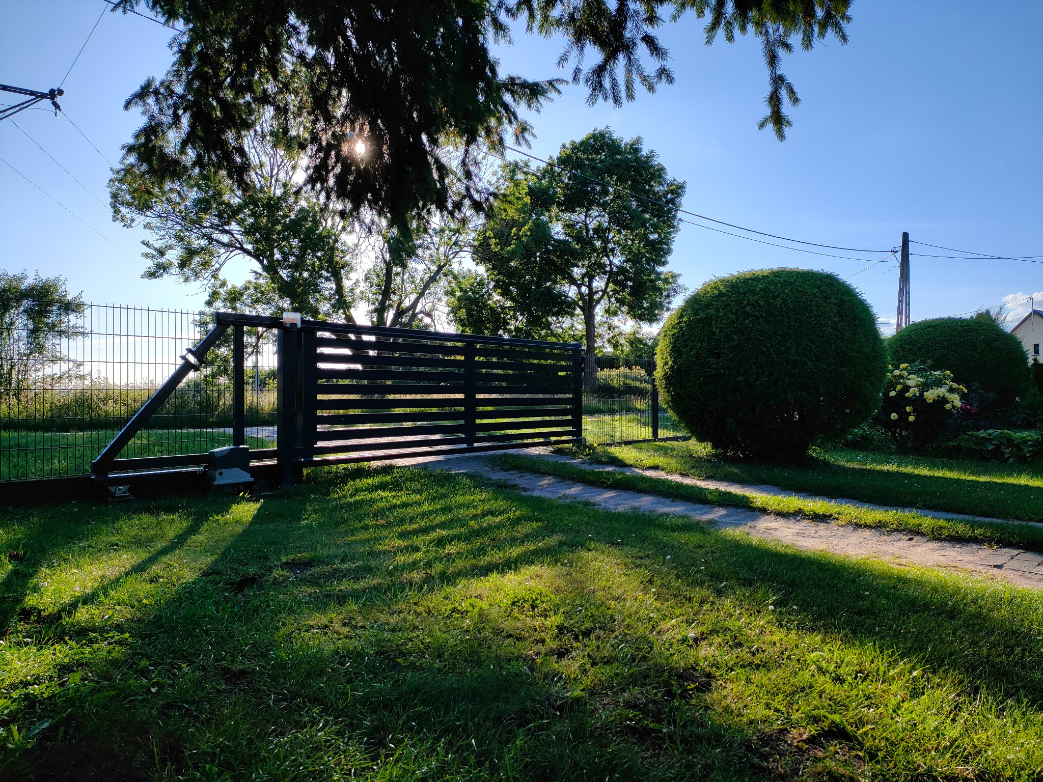 Automatyczna brama wjazdowa z poziomymi panelami w kolorze ciemnego grafitu, widoczna na tle zadbanego trawnika i kulistych krzewów, z promieniami słońca przesączającymi się przez korony drzew.