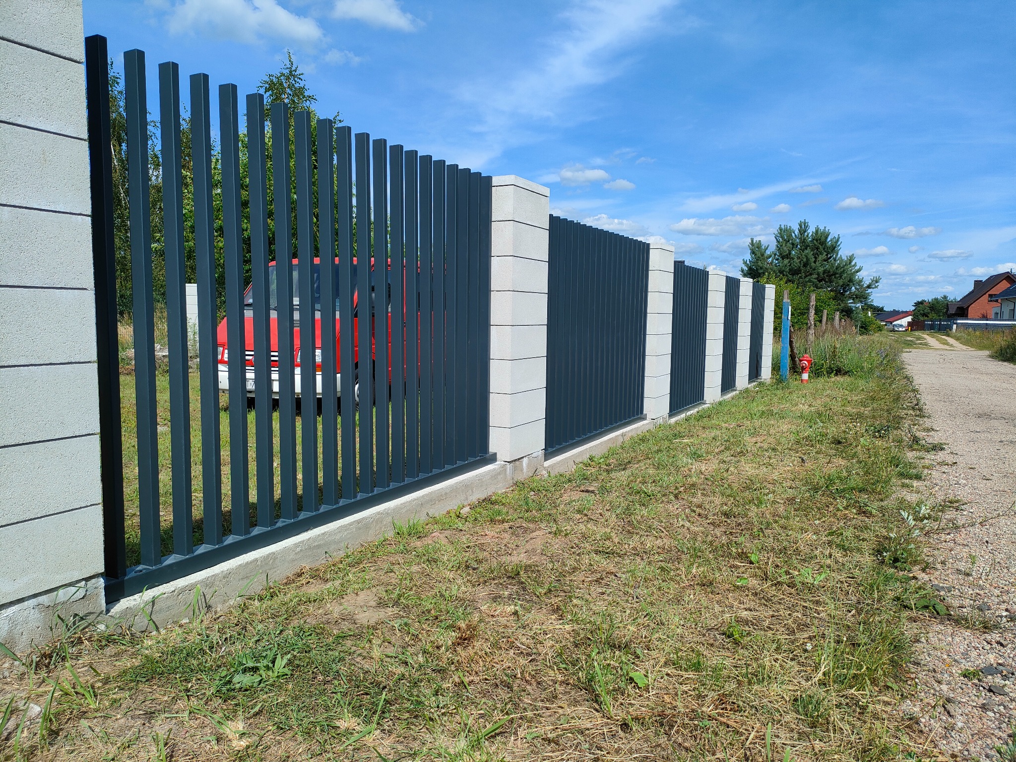 Nowoczesne, grafitowe ogrodzenie z metalowymi sztachetami i betonowymi słupkami, widok z zewnątrz posesji, za ogrodzeniem widoczny czerwony samochód.
