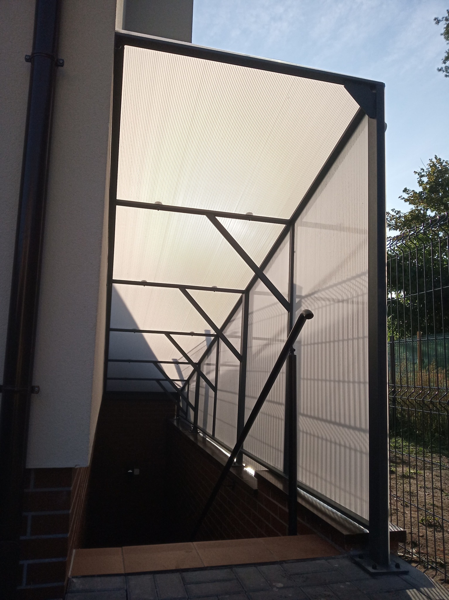 Zadaszone zejście do piwnicy z metalową konstrukcją i wypełnieniem z poliwęglanu, widoczne schody i fragment balustrady, obok ogrodzenie panelowe.