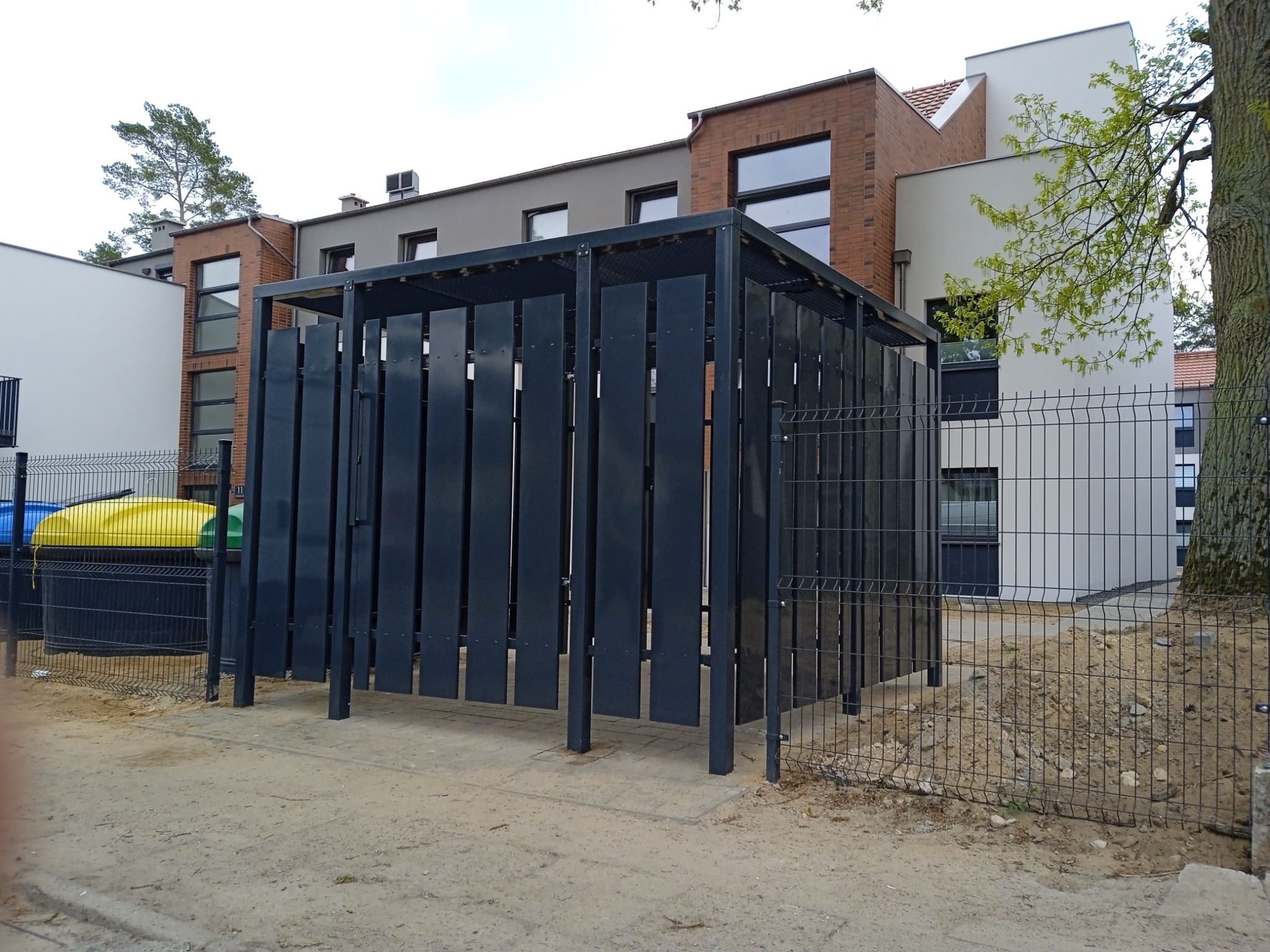 Metalowa konstrukcja osłaniająca pojemniki na śmieci, wykonana z pionowych, ciemnych paneli, stojąca na gruncie, otoczona niskim ogrodzeniem z siatki, na tle nowoczesnych budynków mieszkalnych.