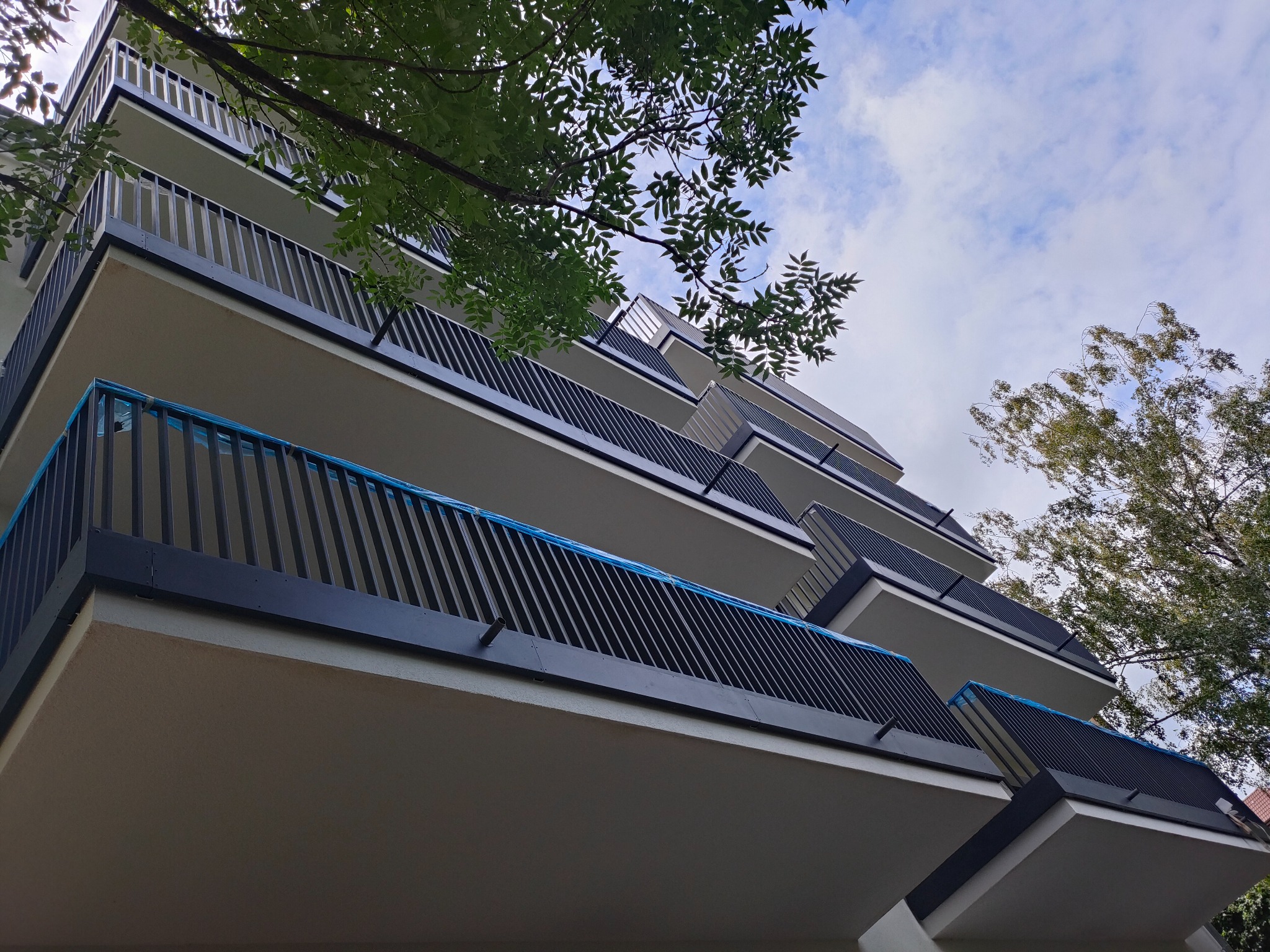 Nowoczesne balkony z czarnymi, metalowymi balustradami w nowym budynku wielorodzinnym, częściowo zabezpieczone folią ochronną, widok z dołu na tle błękitnego nieba z chmurami i zieleni drzew.