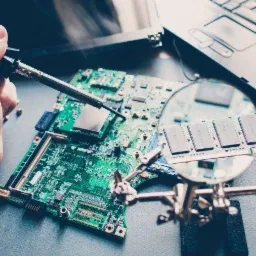 Lutowanie płyty głównej laptopa pod lupą, widoczna kość RAM i elementy elektroniczne, w tle fragment klawiatury laptopa.