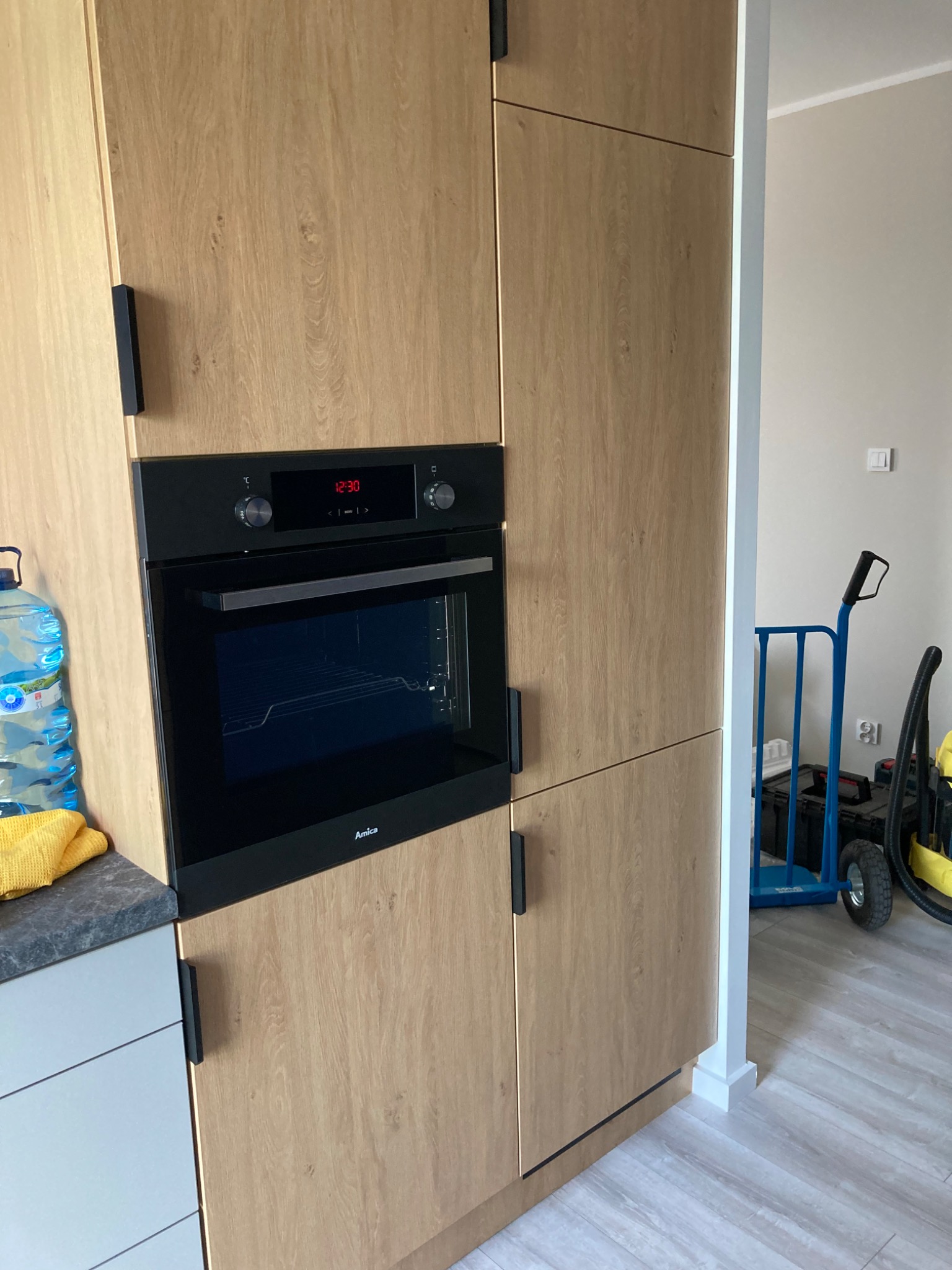Wbudowany piekarnik Amica w zabudowie kuchennej z drewnopodobnymi frontami, widoczny wózek transportowy w tle.
