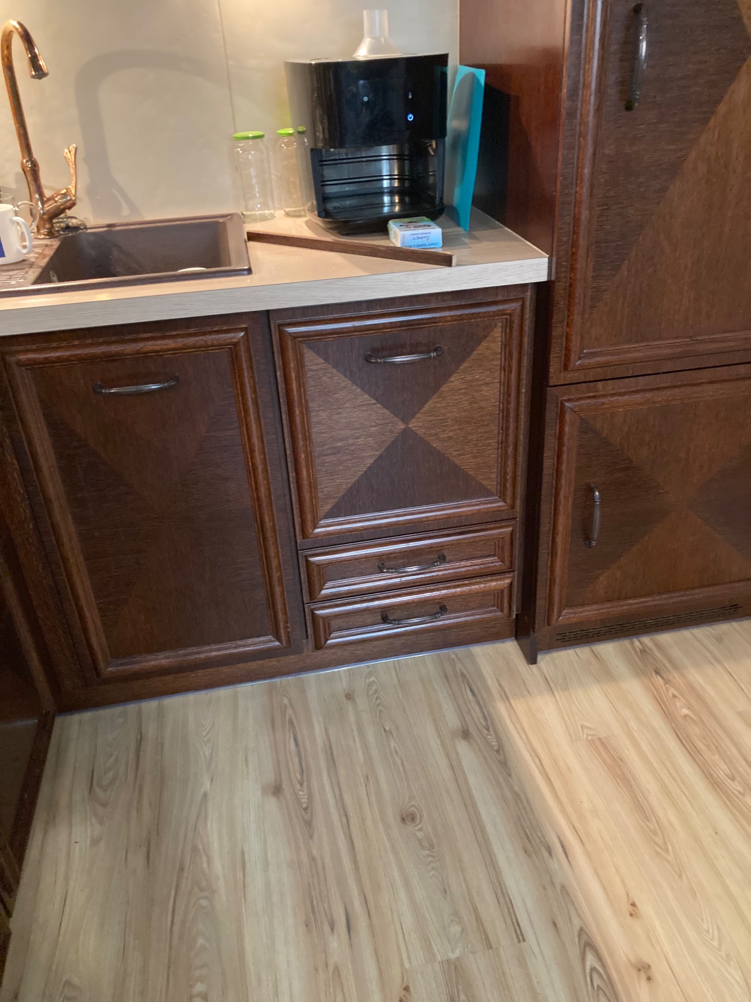 Fragment zabudowy kuchennej z brązowymi, drewnianymi szafkami, blatem, zlewem i baterią, na blacie ekspres do kawy i słoiki, podłoga z paneli.