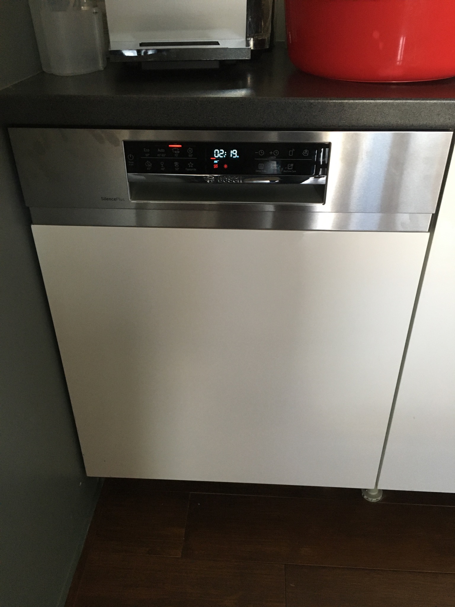 Wbudowana zmywarka marki Bosch z panelem sterowania, na którym widoczny jest wyświetlacz z czasem 02:19, umieszczona w białej zabudowie kuchennej, widoczny fragment podłogi z paneli.