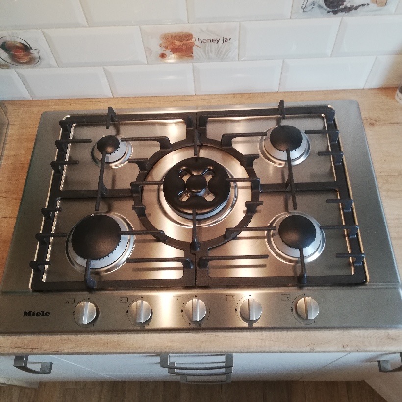 Nowoczesna, czysta płyta gazowa Miele ze srebrnymi pokrętłami i czarnymi palnikami, zamontowana w blacie z drewnopodobnym wykończeniem, widoczna na tle białych płytek z motywami kawy i miodu.