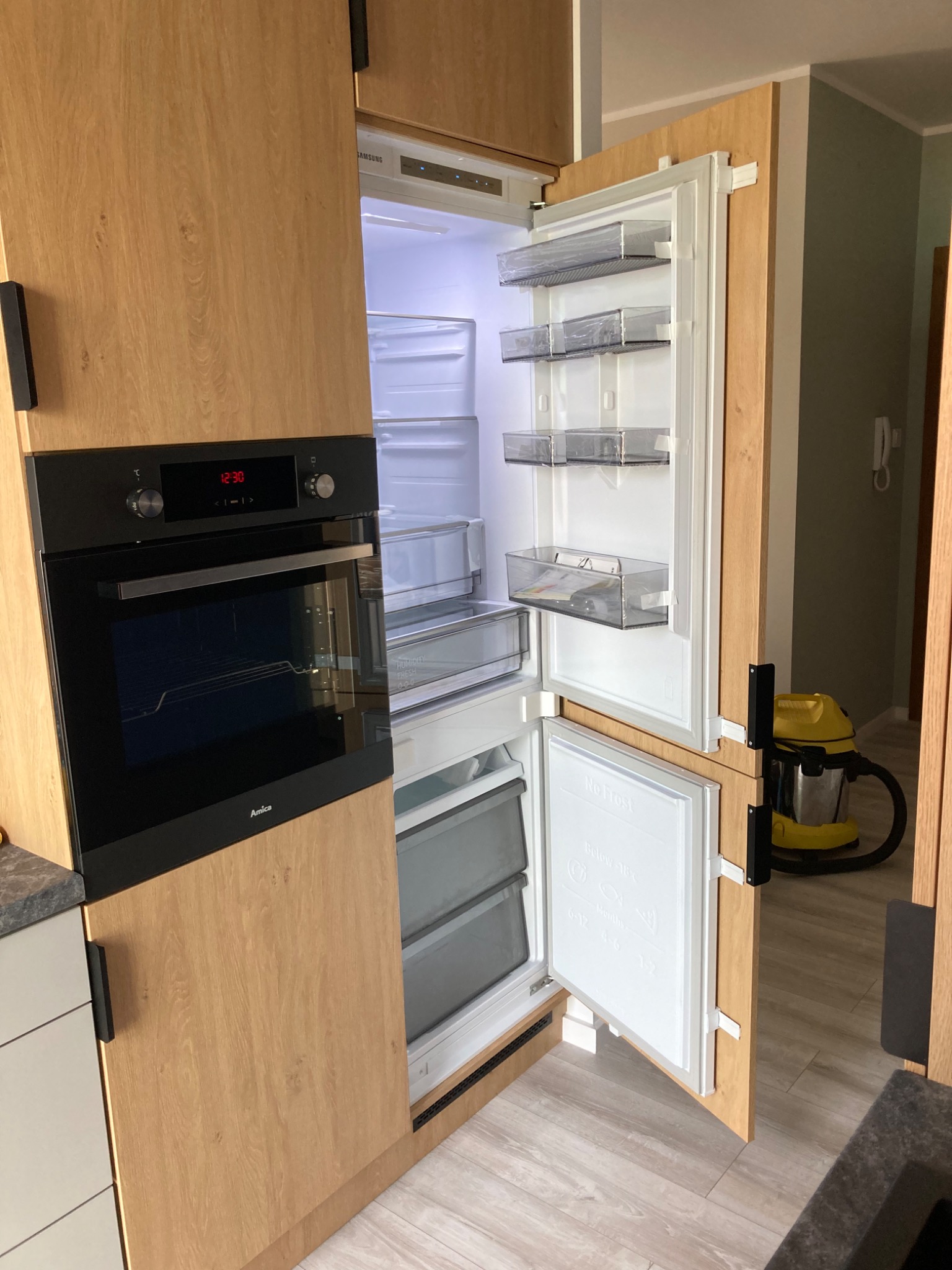 Kuchnia z zabudową meblową w odcieniach drewna, z wbudowanym piekarnikiem Amica i otwartą lodówką Samsung, widoczny odkurzacz w tle.