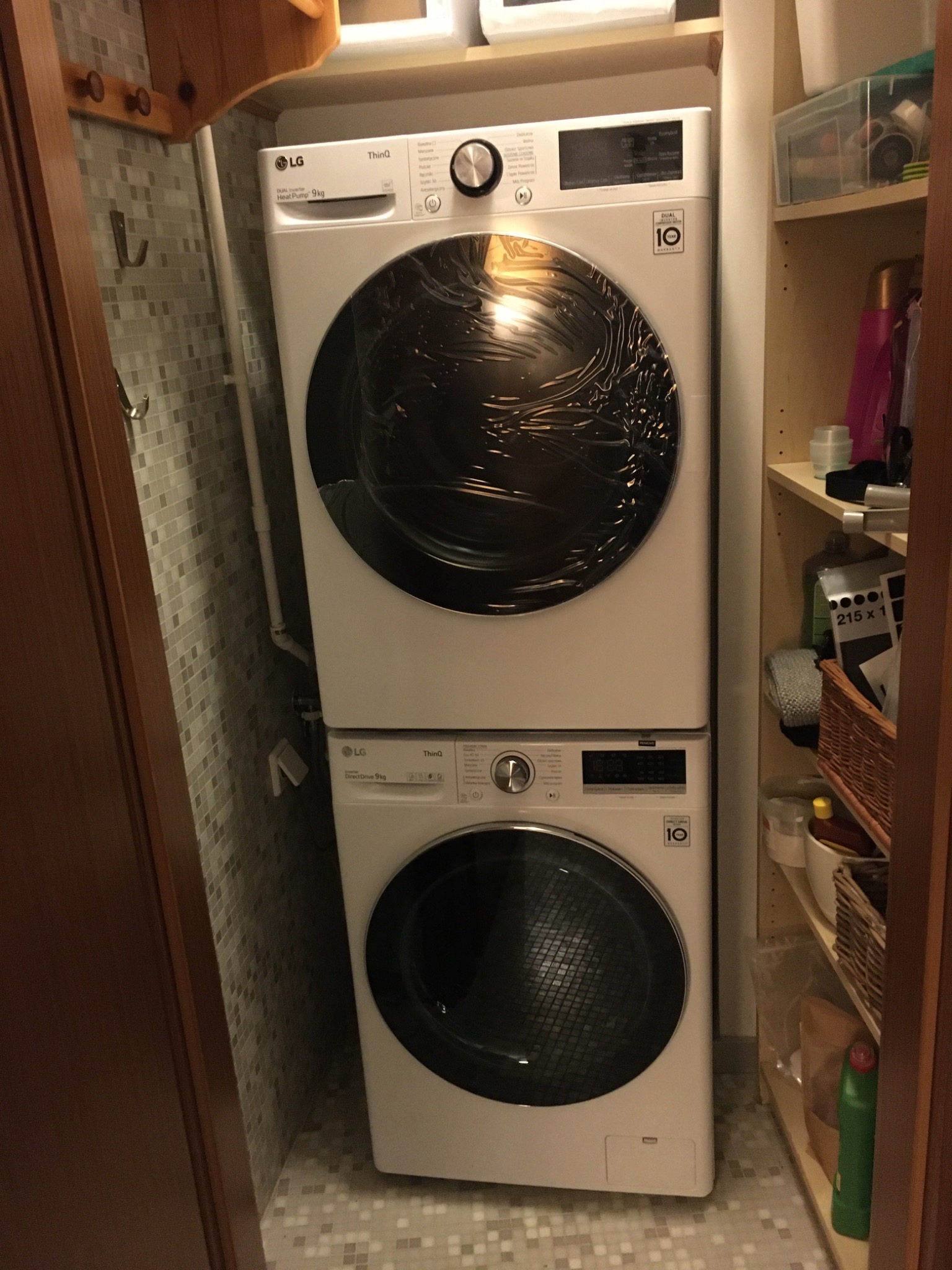 Ustawiony w ciasnej wnęce zestaw pralko-suszarki LG w kolorze białym, z widocznymi panelami sterowania i okrągłymi drzwiczkami, na tle mozaikowej ściany i półek z różnymi przedmiotami.