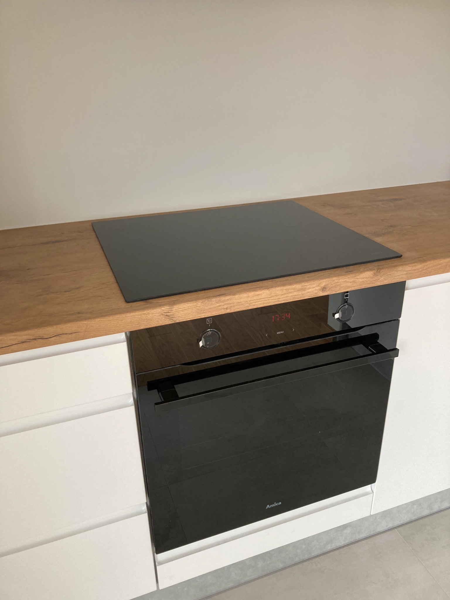 Czarna płyta indukcyjna na drewnianym blacie, zamontowana nad piekarnikiem marki Amica w nowoczesnej, białej kuchni.