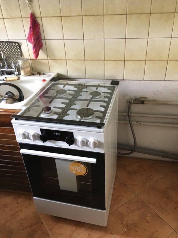 Biała kuchnia gazowo-elektryczna marki Gorenje, z czterema palnikami i szklaną pokrywą, ustawiona w kuchni z beżowymi płytkami, widoczny podłączony przewód gazowy.