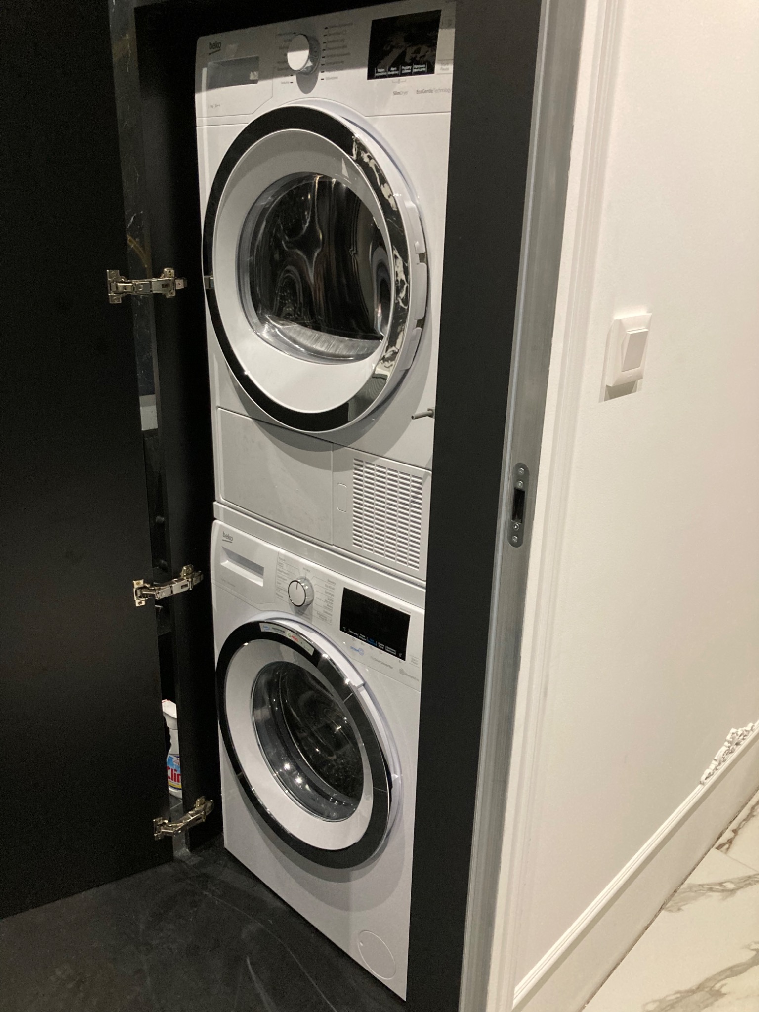 Kompaktowy zestaw pralko-suszarki Beko w zabudowie szafki z otwartymi drzwiami, widoczny fragment marmurowej podłogi i listwy przypodłogowej.