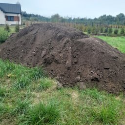 RAFREMS Rafał Cieplak - Dwie pryzmy ciemnej ziemi na trawiastym terenie przed domem jednorodzinnym z szarym dachem w pochmurny dzień.