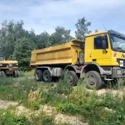 Żółta wywrotka Mercedes Actros i żółta koparka JCB na piaszczystym terenie zarośniętym trawą, w tle widoczne drzewa.