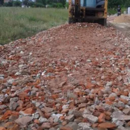Żółta ładowarka czołowa na placu budowy rozgarnia gruz ceglany, widoczne pole i zabudowania w oddali.