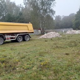 RAFREMS Rafał Cieplak - Żółta wywrotka na łące z kupami gruzu, widoczny fragment lasu i betonowe ogrodzenie w tle, pochmurny dzień.