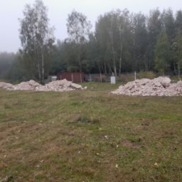 RAFREMS Rafał Cieplak - Żółta wywrotka w oddali, obok trzy pryzmy gruzu na zarośniętym terenie, drzewa w tle, pochmurny dzień.