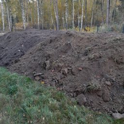 RAFREMS Rafał Cieplak - Kupa ciemnej ziemi z grudami i resztkami roślinności na tle lasu z drzewami liściastymi w jesiennej szacie, widoczne świeże ślady po pracach ziemnych.