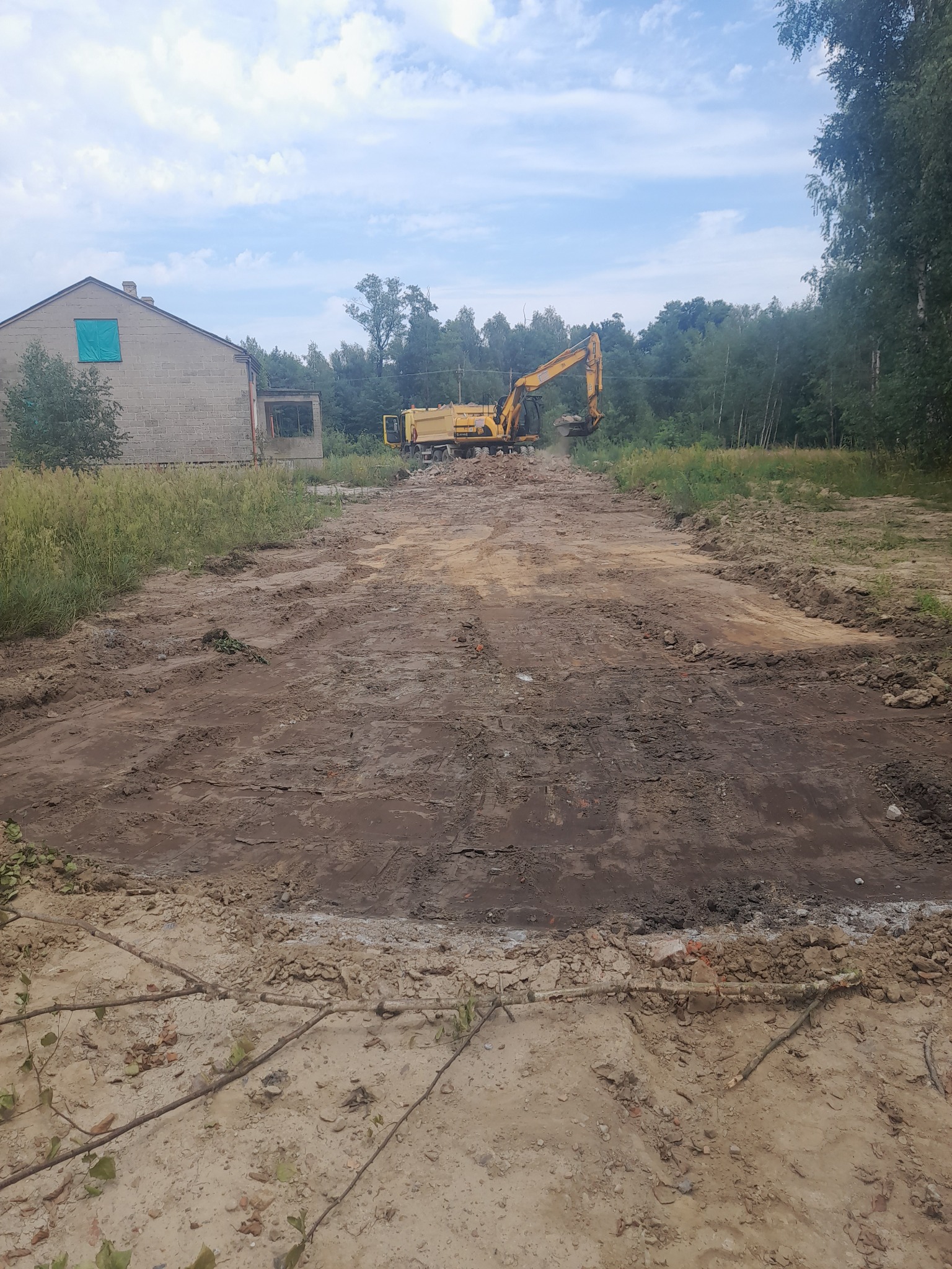 Żółta koparka wyrównuje teren pod budowę w pobliżu opuszczonego budynku z zabitymi oknami, widoczne ślady opon na ubitej ziemi.