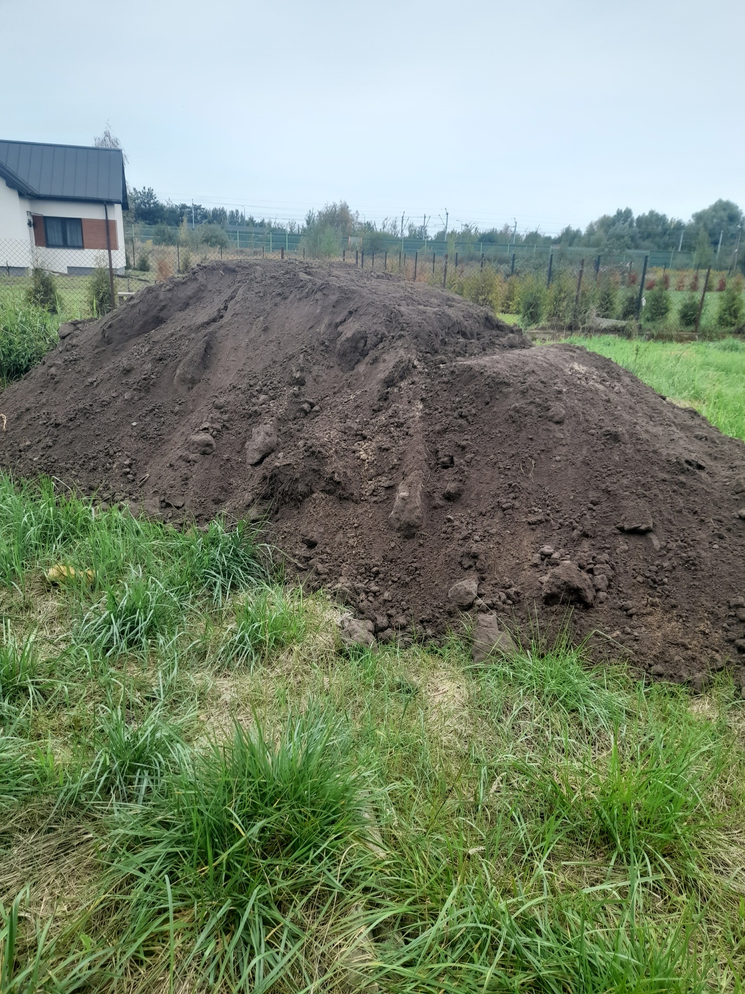 Dwie pryzmy ciemnej ziemi na trawiastym terenie przed domem jednorodzinnym z szarym dachem w pochmurny dzień.