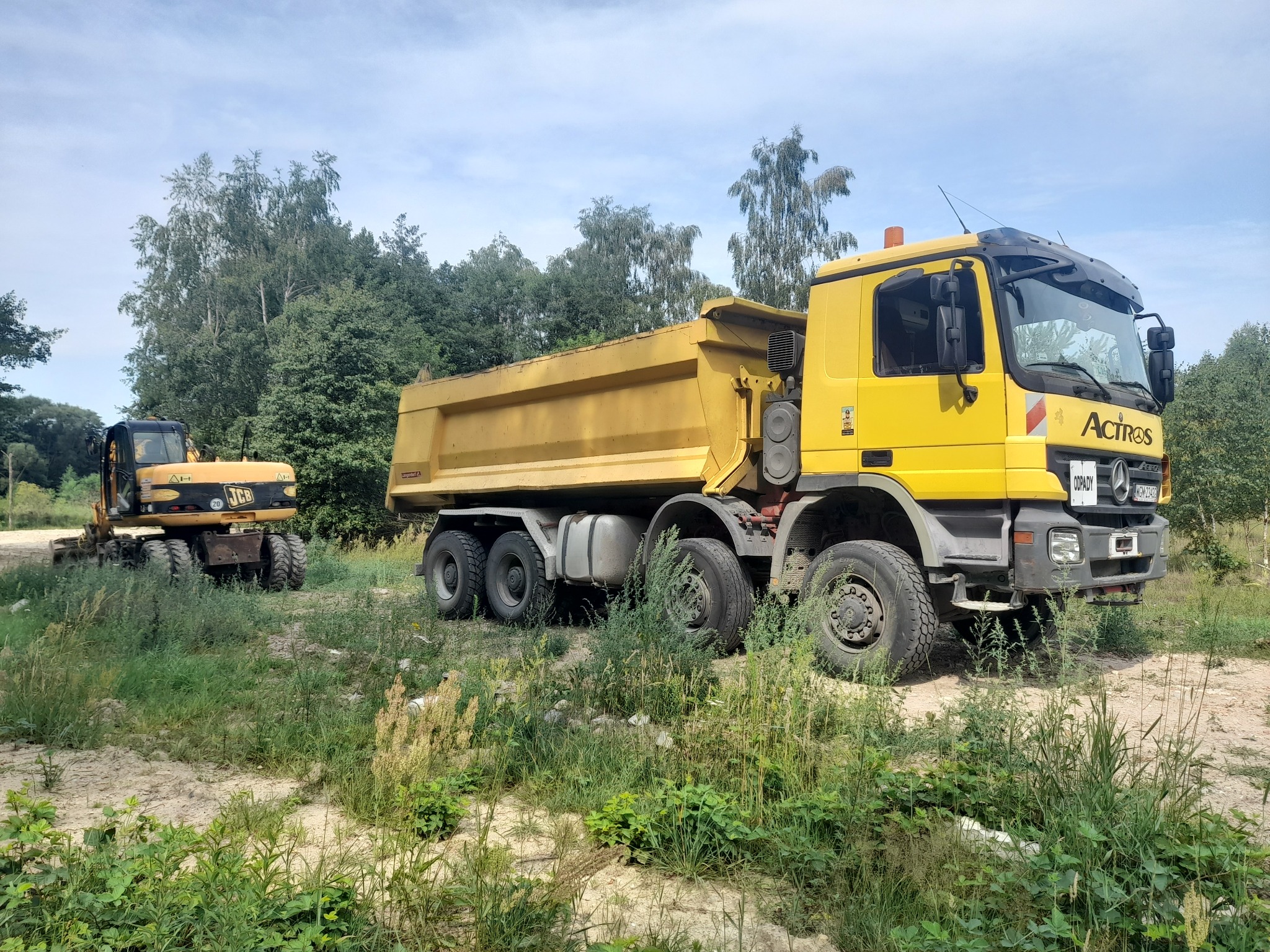 Żółta wywrotka Mercedes Actros i żółta koparka JCB na piaszczystym terenie zarośniętym trawą, w tle widoczne drzewa.