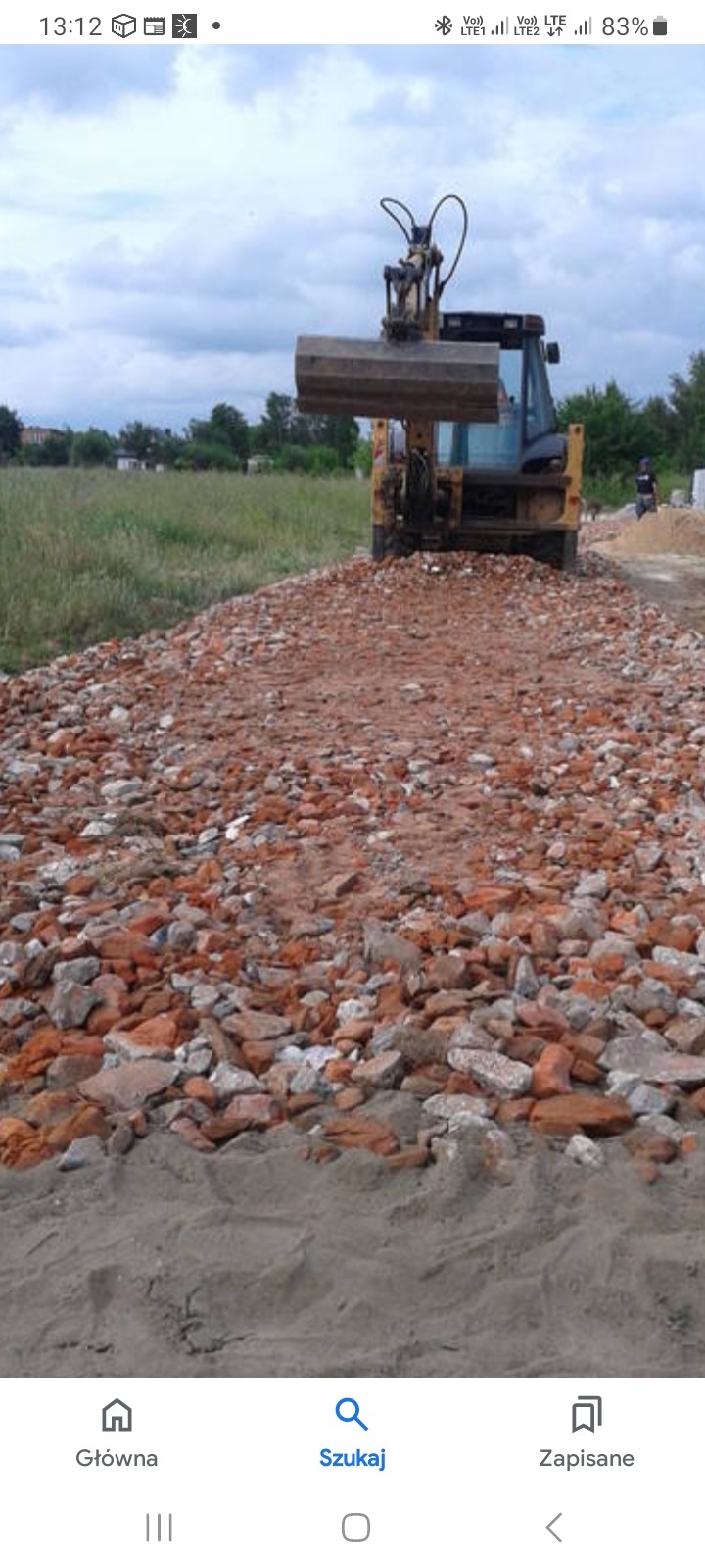 Żółta ładowarka czołowa na placu budowy rozgarnia gruz ceglany, widoczne pole i zabudowania w oddali.