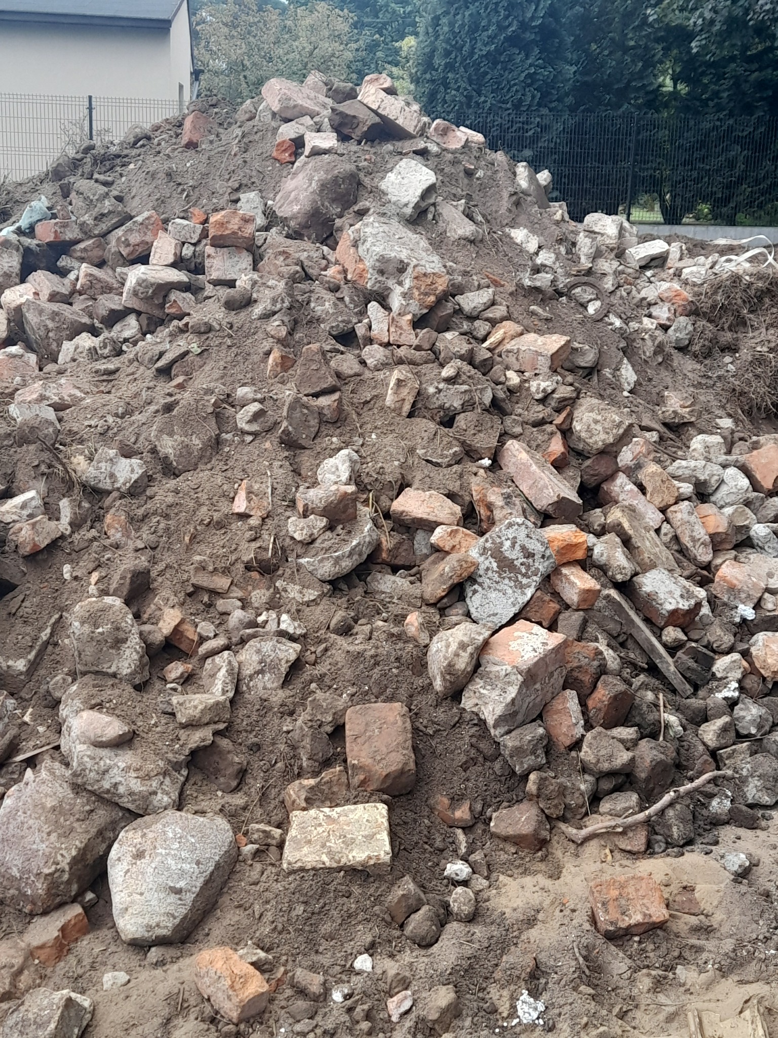 Kupa ziemi zmieszana z gruzem ceglanym i kamieniami na tle ogrodzenia z siatki i zieleni.