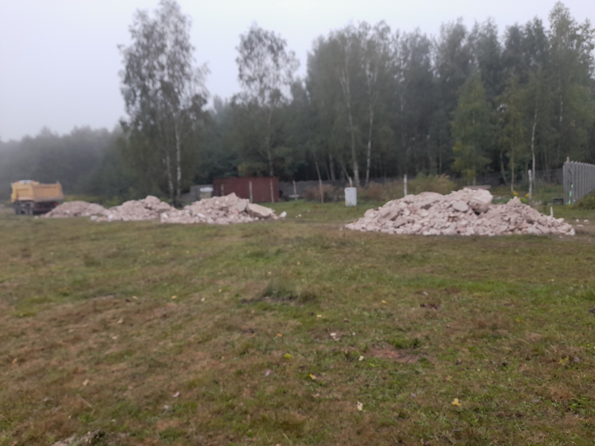Żółta wywrotka w oddali, obok trzy pryzmy gruzu na zarośniętym terenie, drzewa w tle, pochmurny dzień.