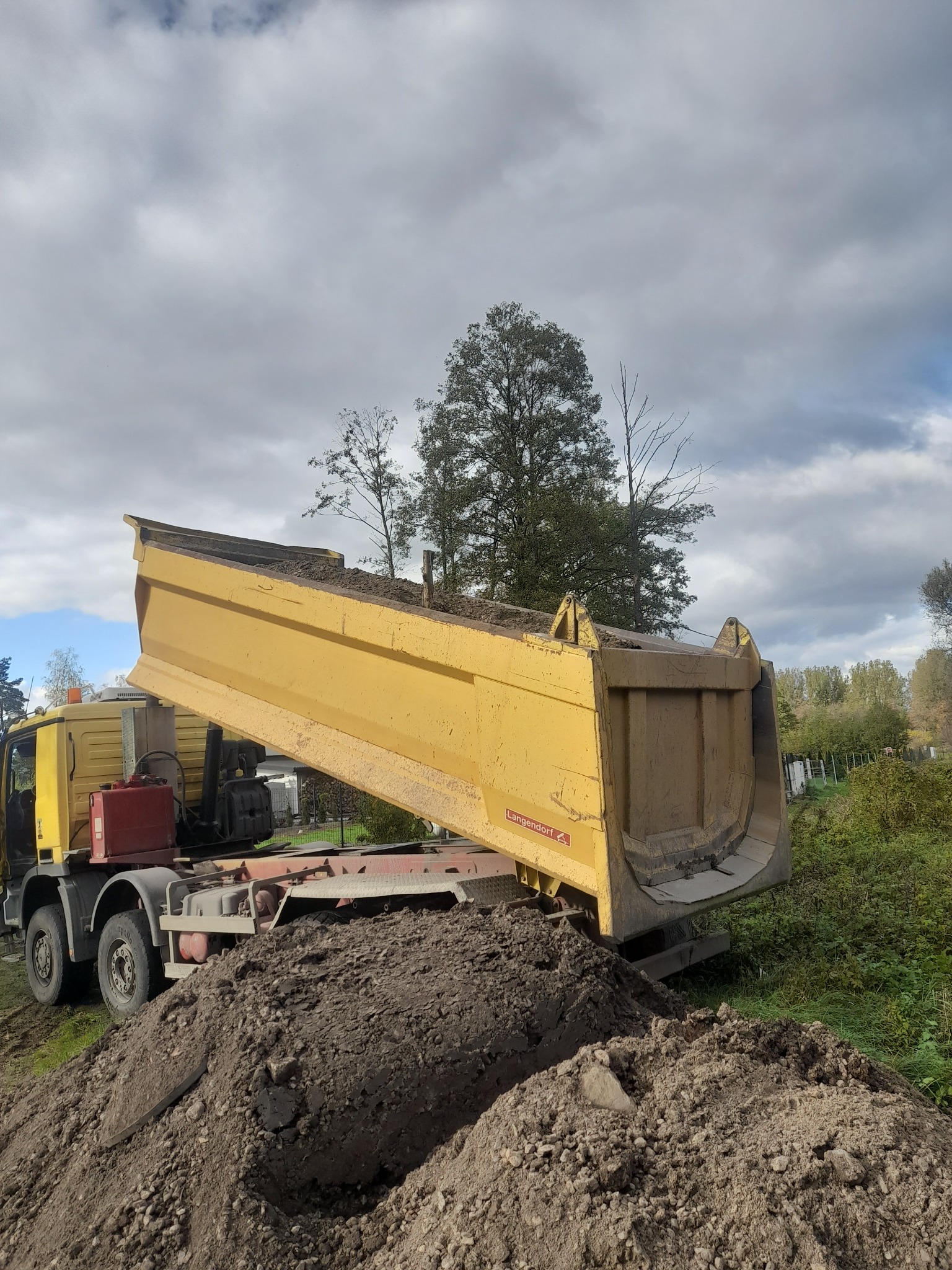 Żółta wywrotka z podniesioną skrzynią ładunkową, wysypująca ziemię na pryzmę, w tle zieleń i pochmurne niebo.