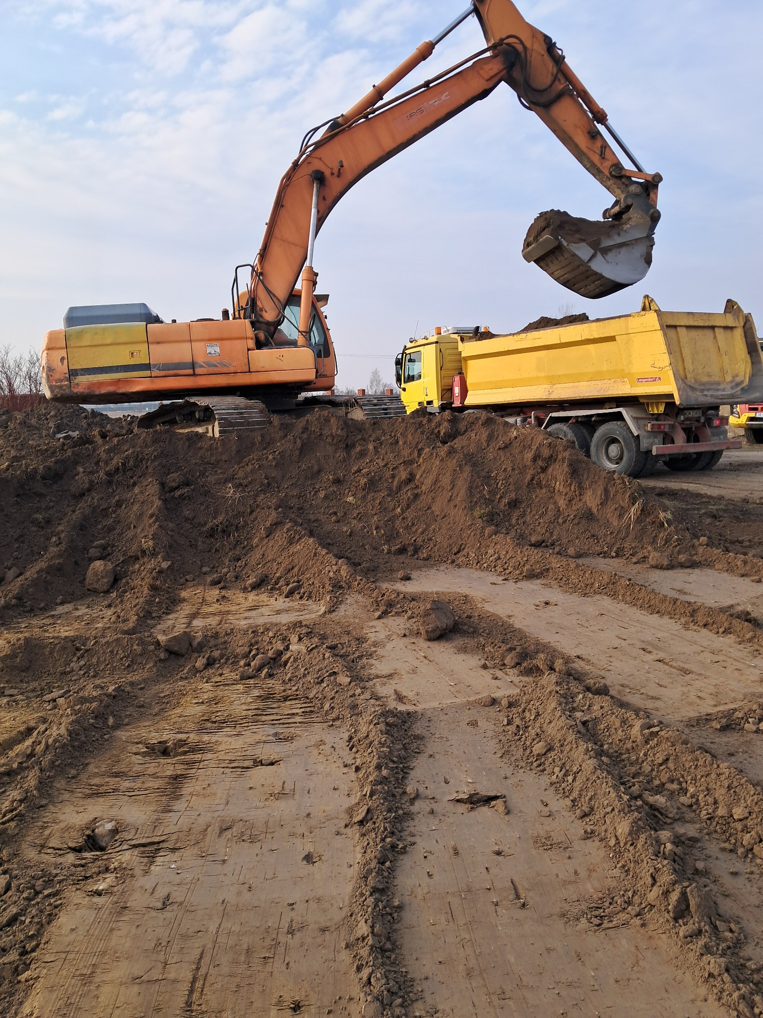 Pomarańczowa koparka nabiera ziemię do żółtej wywrotki na placu budowy, widoczne ślady opon na ziemi.