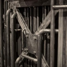 Kuźnia Unikatu Custom Metalwork - Fragment metalowej konstrukcji bramy z elementami rurowymi, z widocznymi spawami i śrubami, w odcieniach szarości.