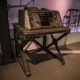 Kuźnia Unikatu Custom Metalwork - Stół warsztatowy z metalową podstawą w kształcie litery X, na którym stoi urządzenie z przezroczystą osłoną, przypominające starą maszynę do pisania lub drukarkę. Tło stanowią ściany o surowym...