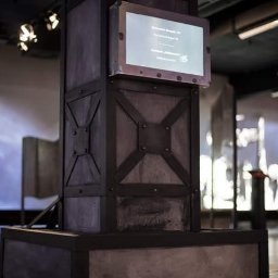 Kuźnia Unikatu Custom Metalwork - Metalowa konstrukcja przypominająca filar z dekoracyjnymi elementami krzyżowymi i ekranem wyświetlającym tekst w muzeum lub galerii.