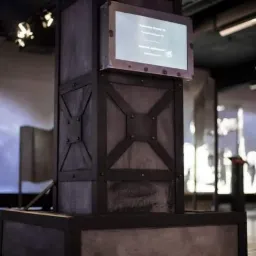 Metalowa konstrukcja przypominająca filar z dekoracyjnymi elementami krzyżowymi i ekranem wyświetlającym tekst w muzeum lub galerii.