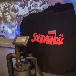 Kuźnia Unikatu Custom Metalwork - Instalacja muzealna: fragment ściany z napisem 'Solidarność' w czerwonej czcionce i flagą Polski, obok metalowa rura z zaworem i ekran dotykowy, w tle zdjęcia z epoki i konstrukcja wieżowa.