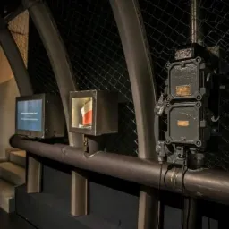 Metalowa instalacja z rurami, skrzynkami i wyświetlaczami na tle siatki, w industrialnym wnętrzu.