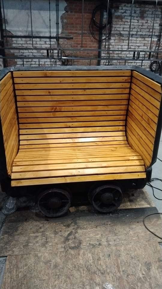 Nietypowa ławka wykonana z drewna i metalu, przypominająca wózek kopalniany, z drewnianymi listwami tworzącymi siedzisko i oparcie, osadzona na metalowych kołach, na tle surowej ściany z cegły...